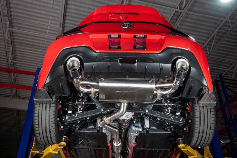 Underbody view of a red Toyota GR86 on a hydraulic shop lift, showing the installed Valvetronic Designs SHUTO valved sport exhaust system with routed mid-pipe, transverse rear muffler, and dual exhaust tips exiting through the rear bumper diffuser.