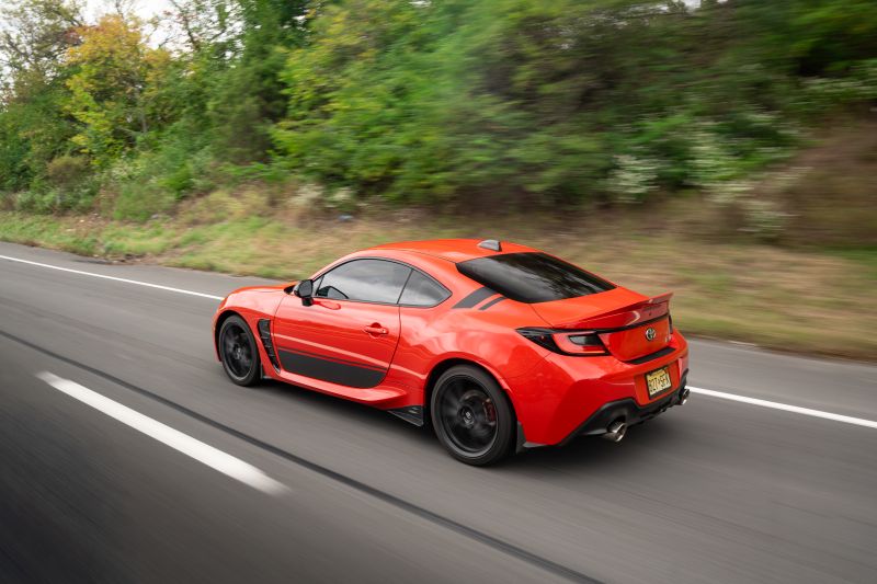 Rear three-quarter rolling shot of a red Toyota GR86 driving at speed on a highway, highlighting the rear diffuser and dual exhaust tips.