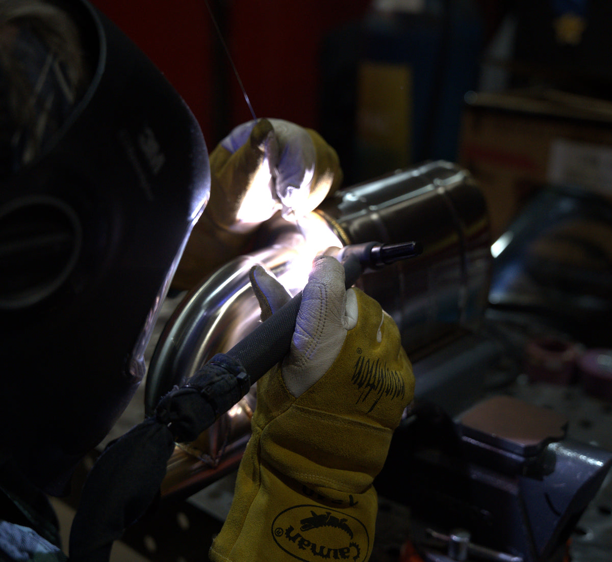Close-up of a metal fabricator TIG welding a performance exhaust muffler, showing the bright welding arc and precision seam work.