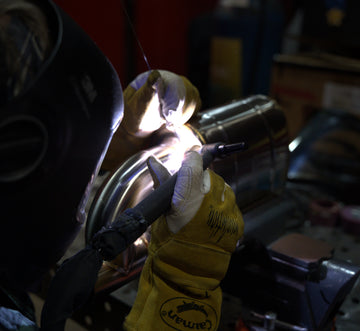 Close-up of a metal fabricator TIG welding a performance exhaust muffler, showing the bright welding arc and precision seam work.