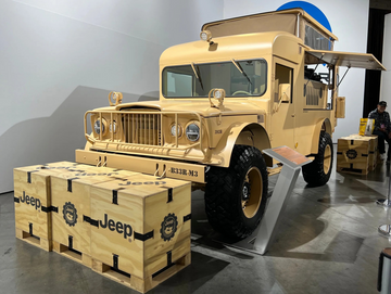Modified desert tan vintage Jeep overlanding concept truck with pop-up roof tent and side-exit exhaust, parked indoors next to wooden "Jeep" crates.