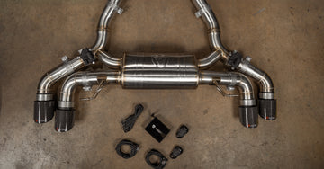 Valvetronic Designs stainless steel valved exhaust system featuring a center muffler, electronic valve actuators, quad carbon fiber exhaust tips, control module box, wiring harnesses, and key fob remotes laid out on a concrete floor.