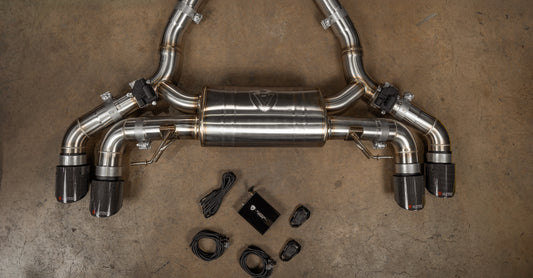 Valvetronic Designs stainless steel valved exhaust system featuring a center muffler, electronic valve actuators, quad carbon fiber exhaust tips, control module box, wiring harnesses, and key fob remotes laid out on a concrete floor.